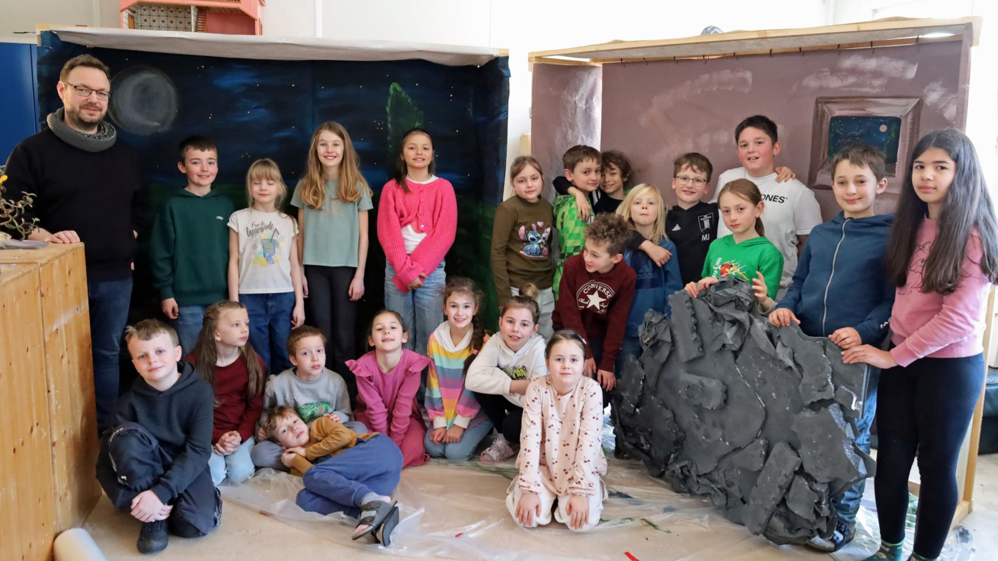 Eine größere Gruppe von Kindern und einer erwachsenen Person steht und sitzt in einem Raum zwischen zwei gebauten Kulissen. Links befindet sich eine Kulisse, die eine nächtliche Landschaft mit Mond und Sternen zeigt. Rechts steht eine zweite Kulisse in warmen Farben mit einem gemalten Fenster. Davor lehnt ein großes künstliches Felsstück. Einige Kinder sitzen auf dem Boden, andere stehen dahinter. Die Gruppe ist umgeben von Holz‑ und Bastelmaterialien, was auf ein gemeinsames kreatives Projekt oder eine Aufführung hindeutet.